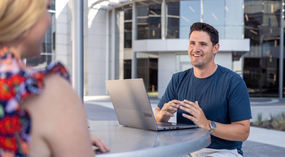 Man sitting in front of open laptop talks to person sitting across the table