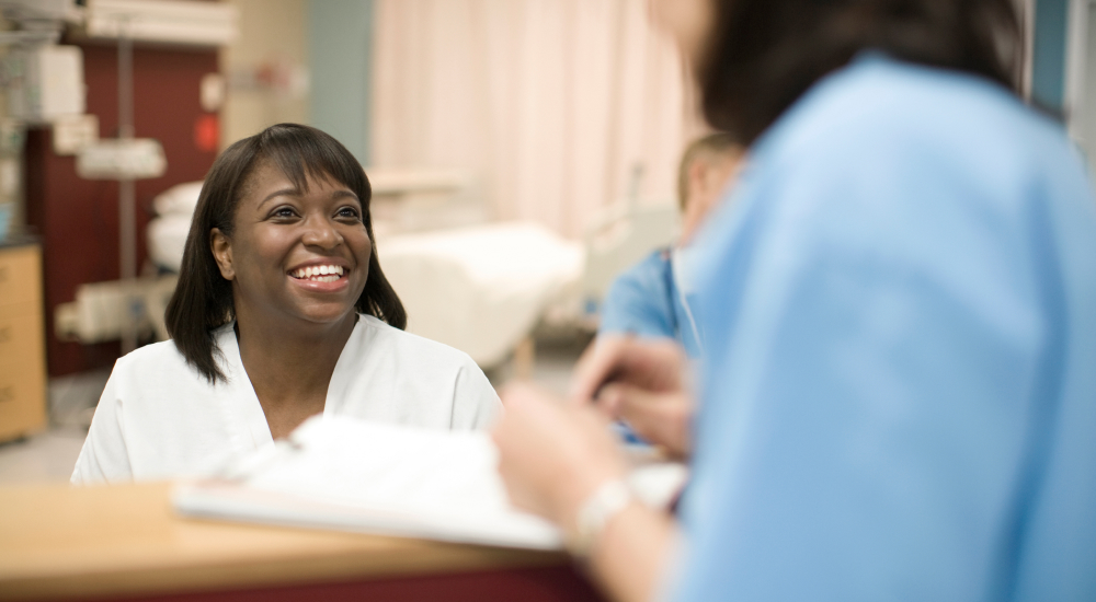 Woman sitting at nurse's desk has conversation with another staff member.