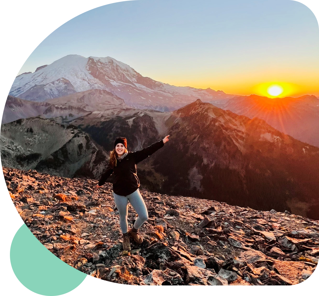A hiker standing on a mountainside at sunset with arms outstretched in triumph.